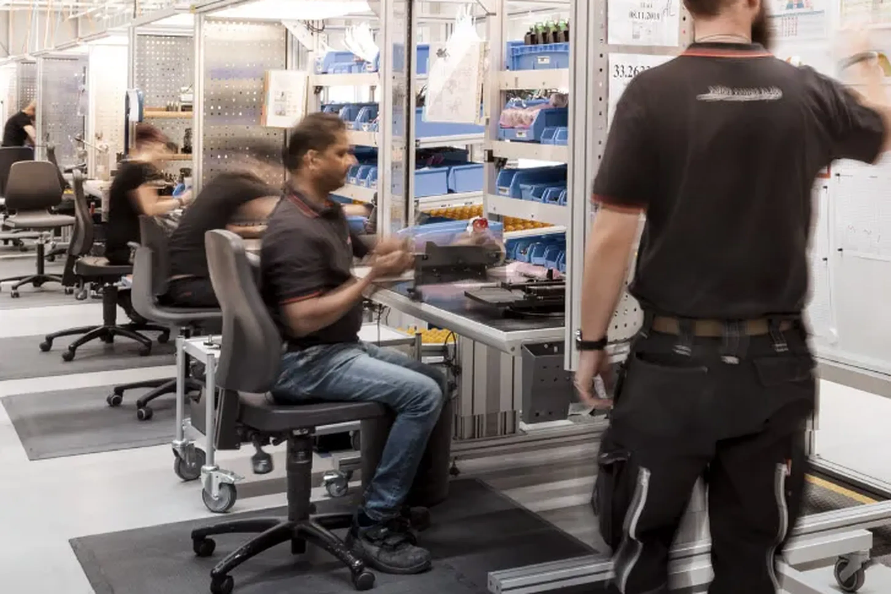 Workers in a modern factory setting, assembling components at workstations with tools and equipment, under bright overhead lighting.