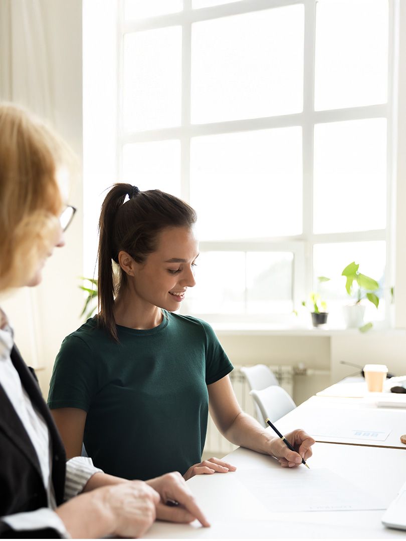 Two people at a table, one writing on paper. Bright room with large windows and plants. Casual and focused atmosphere.