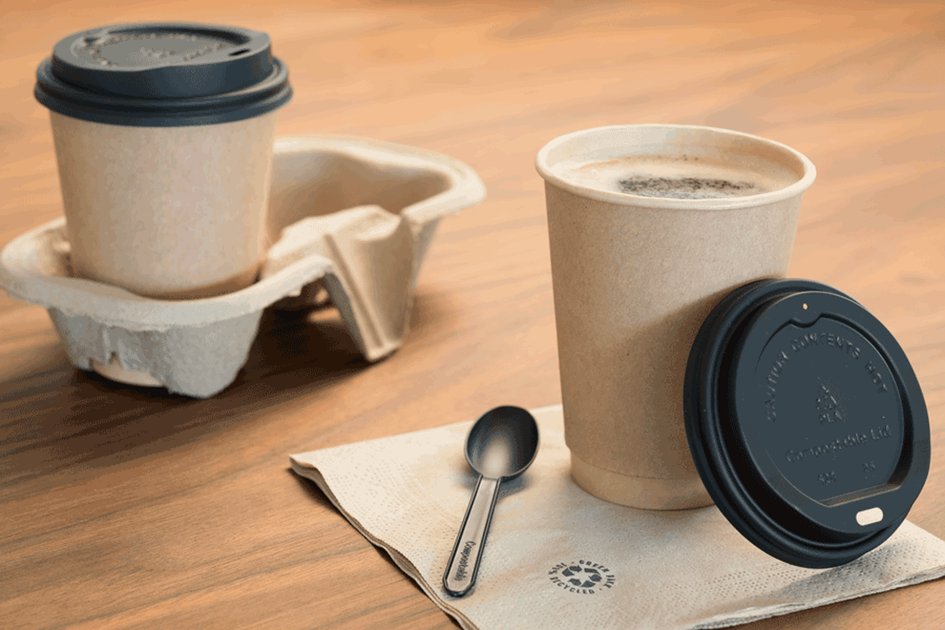 Two brown paper coffee cups with black lids, one on a napkin with a spoon, and the other in a cardboard holder on a wooden table.