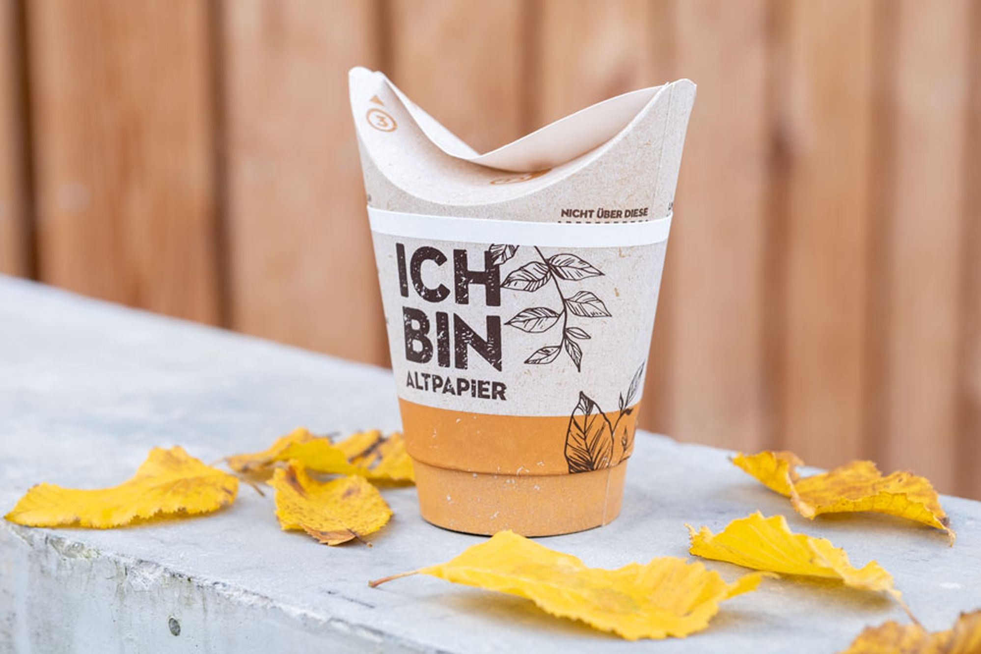 A biodegradable paper cup with "ICH BIN ALTPAPIER" on it, surrounded by yellow leaves on a concrete surface.