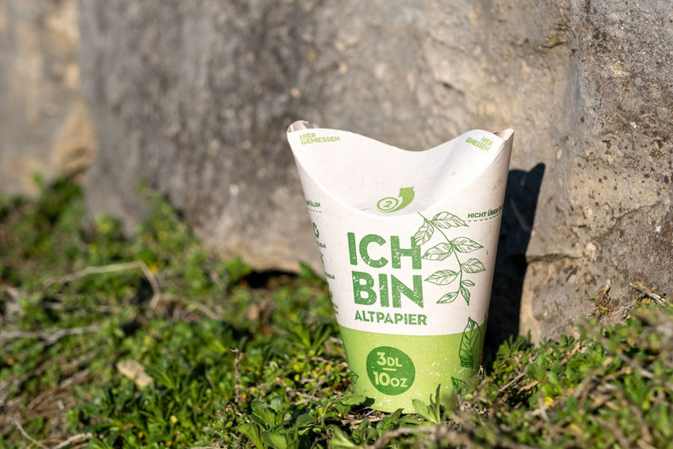 Biodegradable paper cup labeled "Ich bin Altpapier," placed on grass near a stone wall, with green plant graphics.