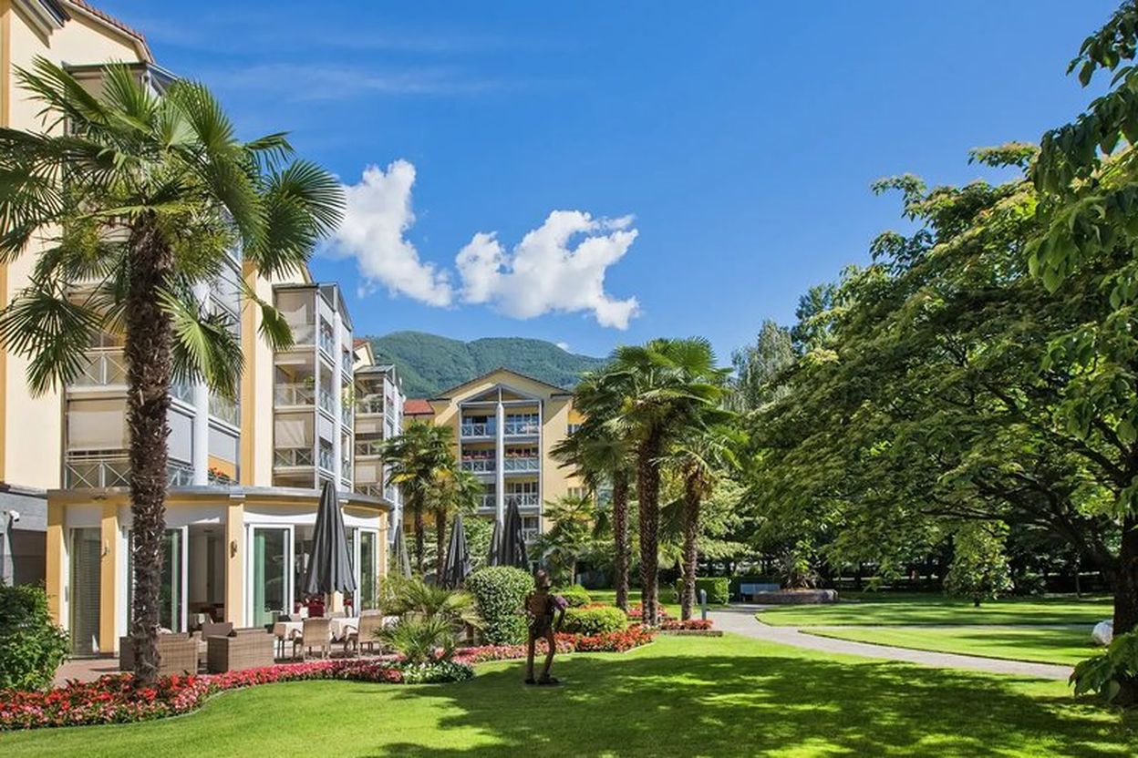 Sunny garden with palm trees and manicured lawns in front of a modern yellow building, set against a backdrop of green hills and a blue sky.