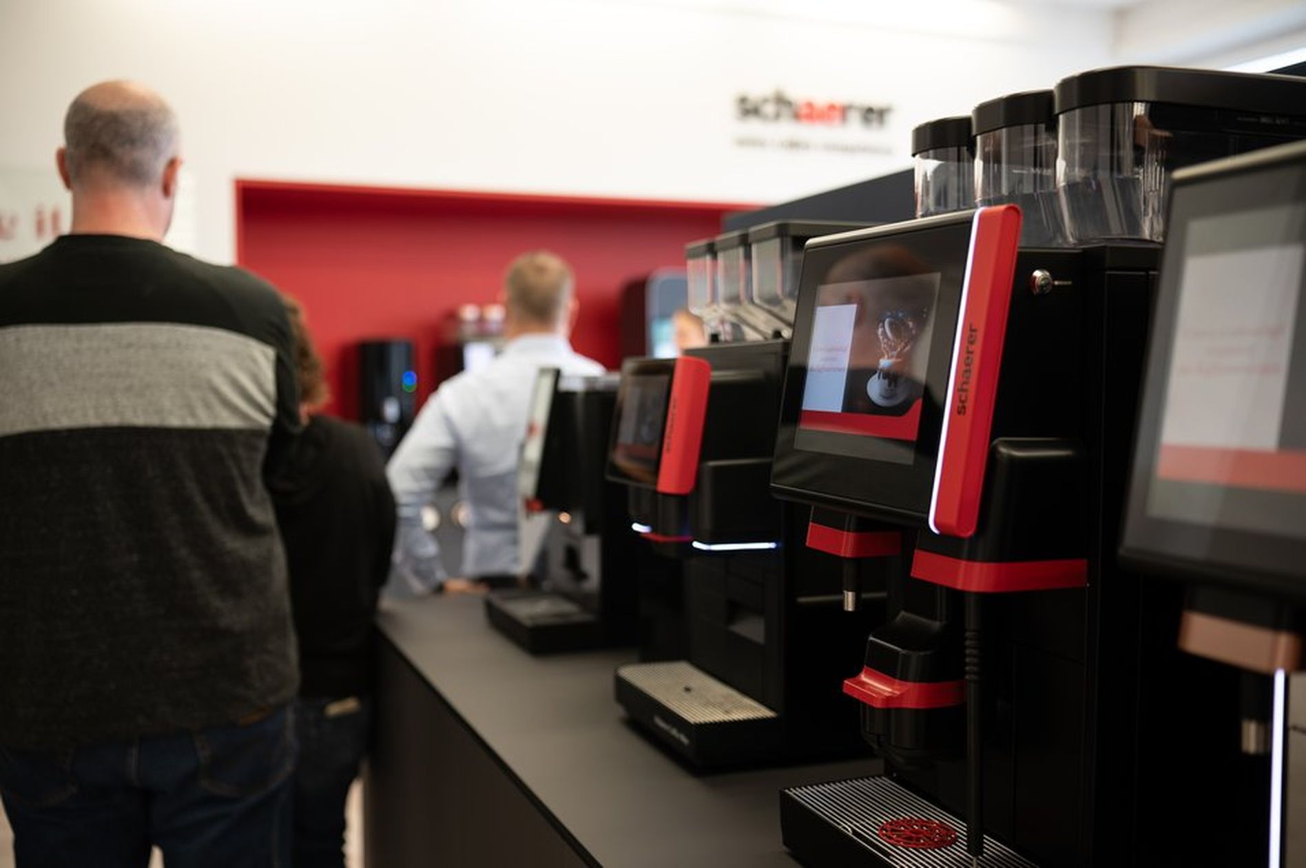 People standing in line at a coffee machine display area, with modern coffee machines featuring digital screens in the foreground.