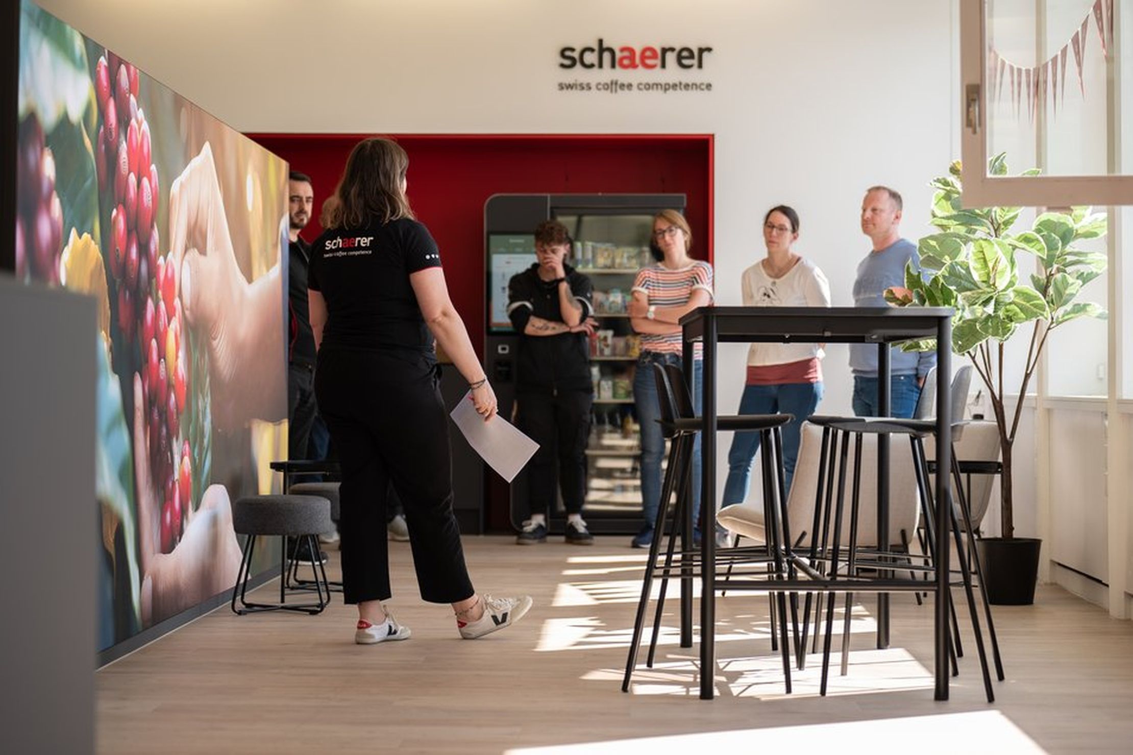 A group of people stands and listens to a presenter in a modern office space with a coffee theme.