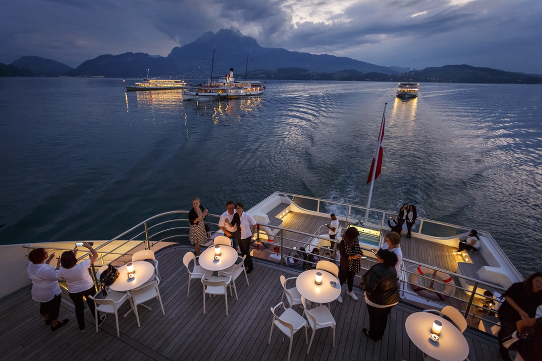People on a boat deck with tables and chairs, enjoying an evening view of a lake and mountains, with other boats in the distance.