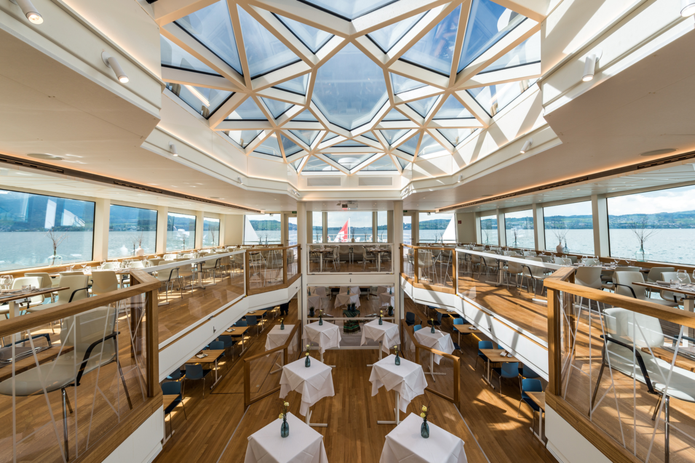 Spacious, modern two-level dining area with large geometric skylight, wooden floors, and lake views through wide windows. Tables set with white cloths.