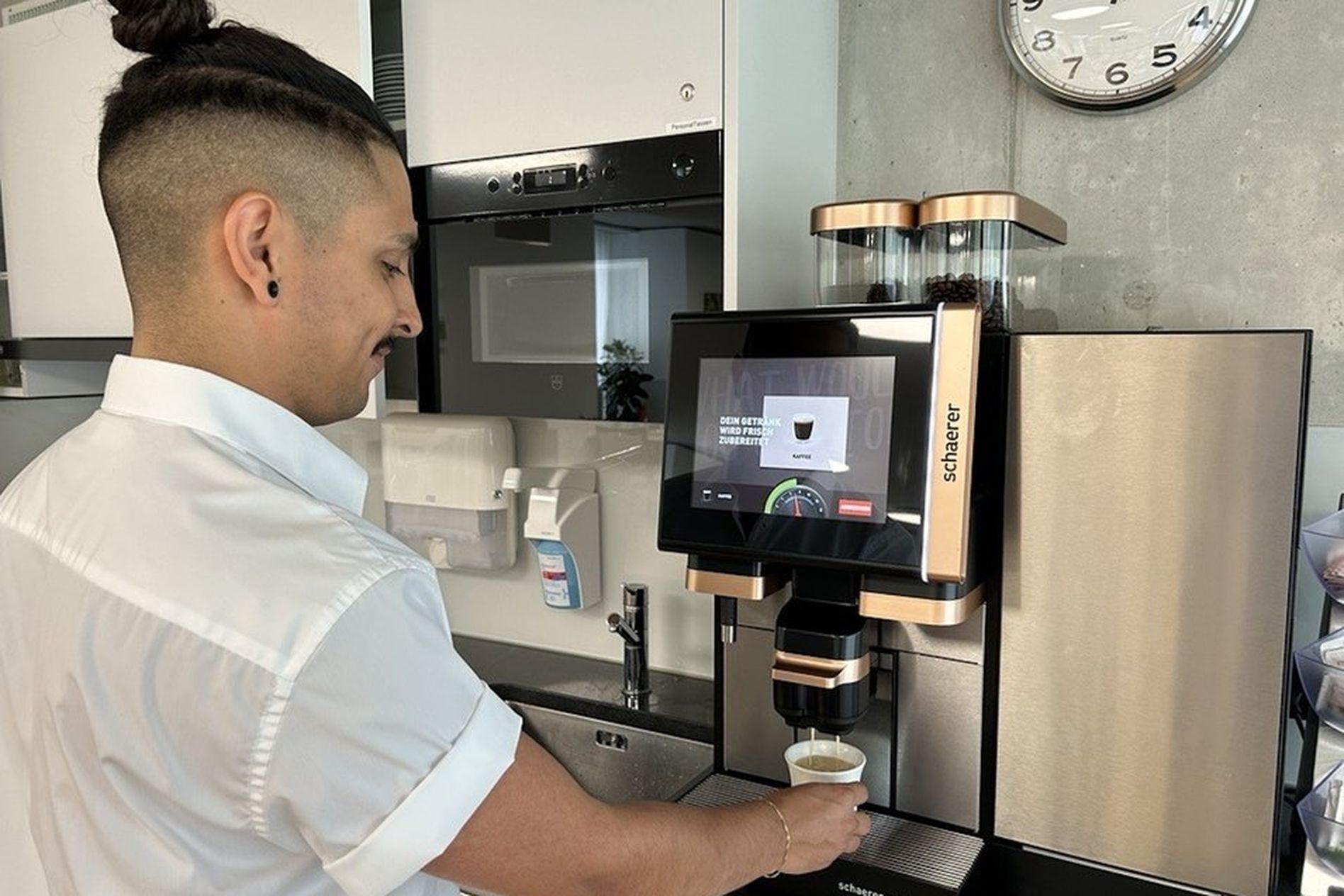 Person using a modern coffee machine in a kitchen, filling a cup. A clock on the wall shows 10:10.